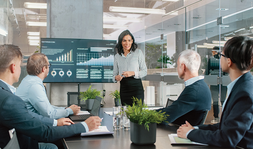 Woman stands at the head of a table and presents to a group of man in suits.