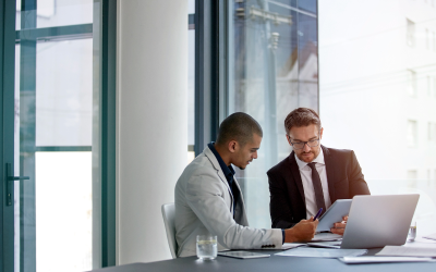 Two HR professionals sit at a table in a modern office. They are focused on a tablet and an open laptop, discussing strategies for managing layoffs and the benefits of outplacement.