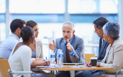 The image depicts a group of individuals seated around a table in a business meeting. The group of six people are fully engaged in the meeting, suggesting a collaborative space. The individual in the middle leads the discussion. The table has papers and bottles of water, indicating an office setting.