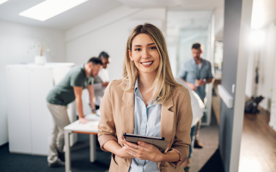 Identifying high potential employees for leadership roles can be challenging. This image shows a high potential future leader, identified through leadership assessments, confidently holding a tablet in an office environment with employees working in the background.