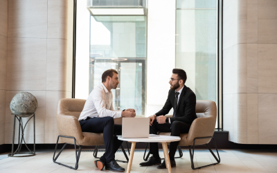 A coach and a departing senior leader sit across from each other in a one-on-one session, discussing goals and next steps as part of outplacement services for executives. A laptop and a small table sit between them. 