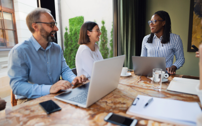 Three engaged employees collaborate at a conference table in a modern office, with two open laptops in use. Their focused discussion reflects the positive impact of their organization's comprehensive career management strategy on employee engagement.