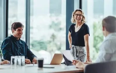 A confident leader stands at the head of a conference table, surrounded by engaged, smiling employees with a laptop and documents in front of them. After facing challenges with engagement and retention, the company invested in leadership coaching to help answer the question: how to retain top talent — and saw positive results across the organization.