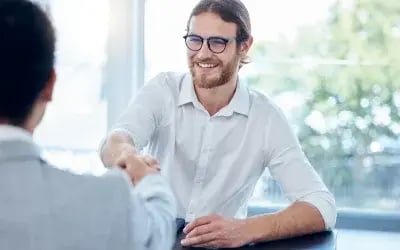 A person wearing glasses and a button-down shirt smiles and shakes hands with the hiring manager after a successful interview, thanks to the expert job interview tips they found online.