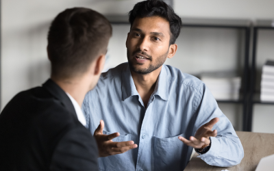 A coach and coachee sit at a table in a modern office, engaging in Right Management's leadership coaching services. Office shelving lines the background.