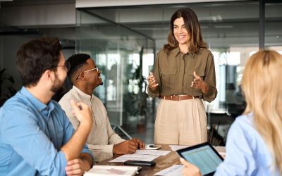 A business leader stands at the head of a conference table, guiding a successful meeting with three fully engaged employees. The modern office setting, with papers, notebooks and a tablet on the table, highlights a culture of collaboration and strategic thinking. This scene reflects the impact of a strong, unbiased hiring decision made through leadership assessments.