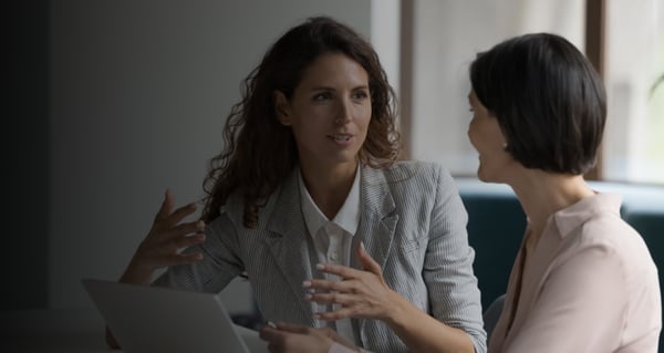 A career coach and a coachee, both dressed in professional business attire, sit at a conference table with a laptop open between them, actively engaging in a one-on-one coaching session as part of an outplacement program.