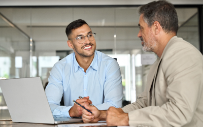 A coach and a coachee are seated at a table in a modern office, with a laptop open in front of them. They are engaged in a discussion about the coachee's future career decisions. This coaching session is part of the outplacement service provided by the coachee's former employer.